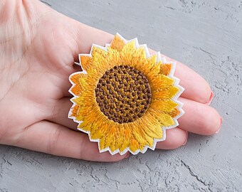 Pode incluir: Um remendo de girassol bordado amarelo com um centro marrom. O remendo está em um fundo branco.