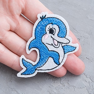 Puede incluir: Un parche bordado de un delfín sonriente, con colores azul y blanco y un fondo blanco.