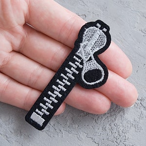 Puede incluir: Un parche de hierro bordado en blanco y negro de una cremallera con una lengüeta. La cremallera es blanca y la lengüeta es negra.