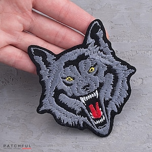May include: Embroidered wolf patch with gray fur, yellow eyes, and a red open mouth. The patch has a black border and is designed for application on clothing or accessories. The patch is a detailed applique.
