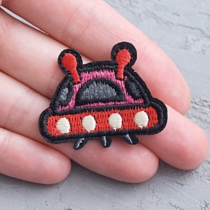 Può includere: Una toppa ricamata che rappresenta un'astronave di cartoni animati in rosso e nero con oblò bianchi.