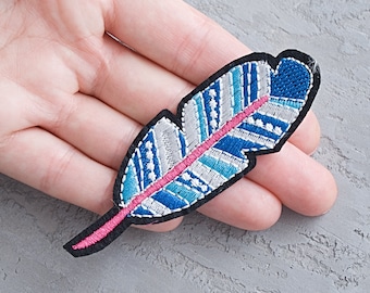 May include: An embroidered feather patch in blue, white and pink. The feather is detailed with a white and blue striped pattern and a pink centre line.