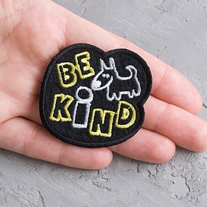 Puede incluir: Parche bordado negro con las palabras "BE KIND" en amarillo y blanco, junto con una ilustración de un perro de dibujos animados. El parche tiene forma redondeada.