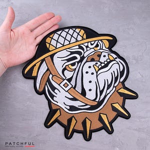 May include: Embroidered patch featuring a bulldog wearing a hat and spiked collar. The patch has a black border, with white, tan, and yellow accents. The bulldog's face is detailed with black outlines and yellow eyes.