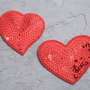 May include: Two red sequin hearts on a grey textured background.