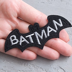 以下が含まれることがあります： 黒いフェルトのアイロン接着パッチで、白い刺繍の蝙蝠のシルエットと、その中に「BATMAN」の文字が入っています。