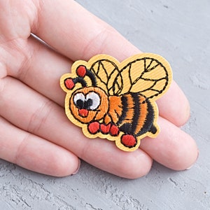 May include: A yellow embroidered bee patch with black stripes, orange legs, and white eyes. The bee is smiling and has a red tongue.