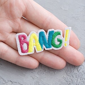 May include: A white fabric patch with the word "BANG!" embroidered in pink, yellow, blue, and green.
