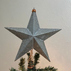 May include: A gray star-shaped Christmas tree topper with a copper wire spring attached to the bottom. The star is made of a textured material and has a rustic look.