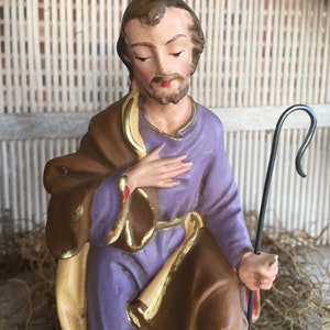 May include: A vintage figurine of a kneeling man wearing a purple robe and brown cloak, holding a staff. The figurine is made of painted plaster and has a rustic, aged appearance.