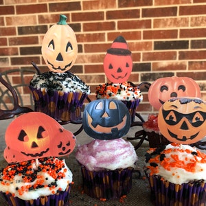 May include: Six cupcakes decorated with Halloween-themed pumpkin toppers. The cupcakes have white frosting and orange and black sprinkles. The pumpkin toppers are in various colors and designs.
