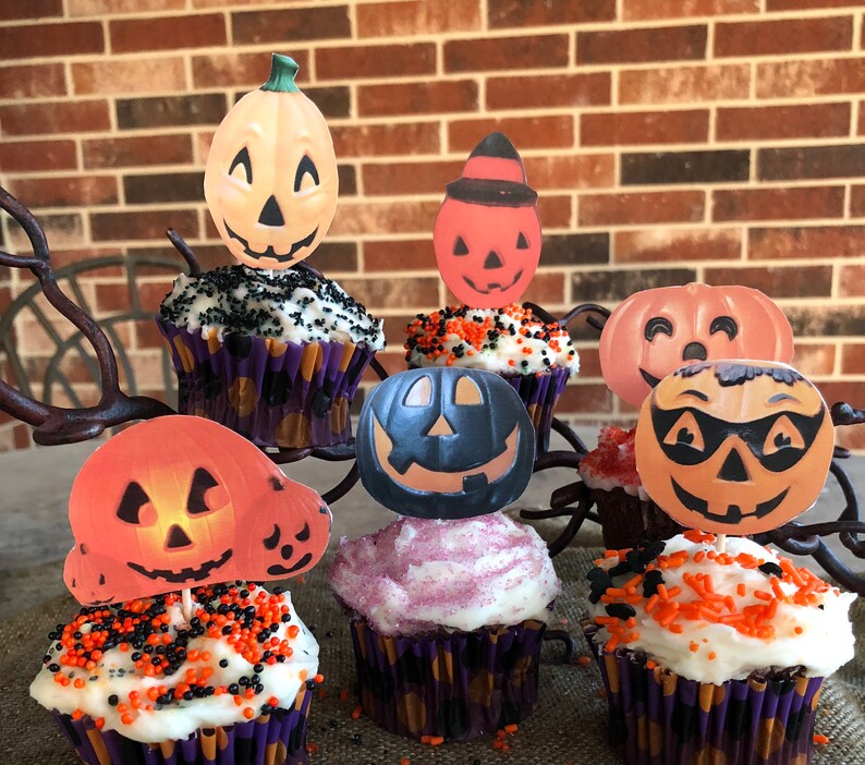 May include: Six cupcakes decorated with Halloween-themed pumpkin toppers. The cupcakes have white frosting and orange and black sprinkles. The pumpkin toppers are in various colors and designs.