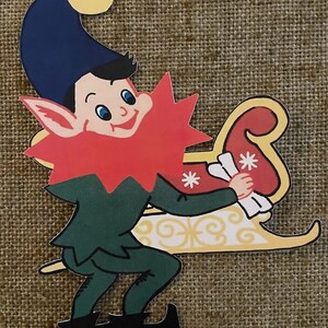 May include: A green and red elf with a blue hat and a yellow pom-pom sits in a yellow sleigh with white snowflakes. The elf is holding a white scroll.