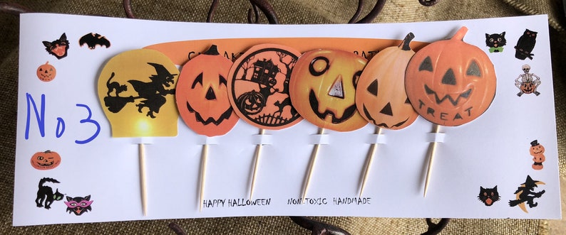 May include: A set of six Halloween-themed cupcake toppers featuring orange and black jack-o'-lantern faces, a witch flying on a broomstick, a haunted house, and the word "Treat". The toppers are on wooden skewers and are labeled "No. 3".