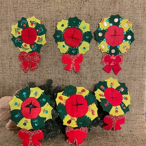 May include: Six felt Christmas wreaths with green, yellow, and red accents. Each wreath has a red center with a white cross and is decorated with sequins and beads. The wreaths have red bows.