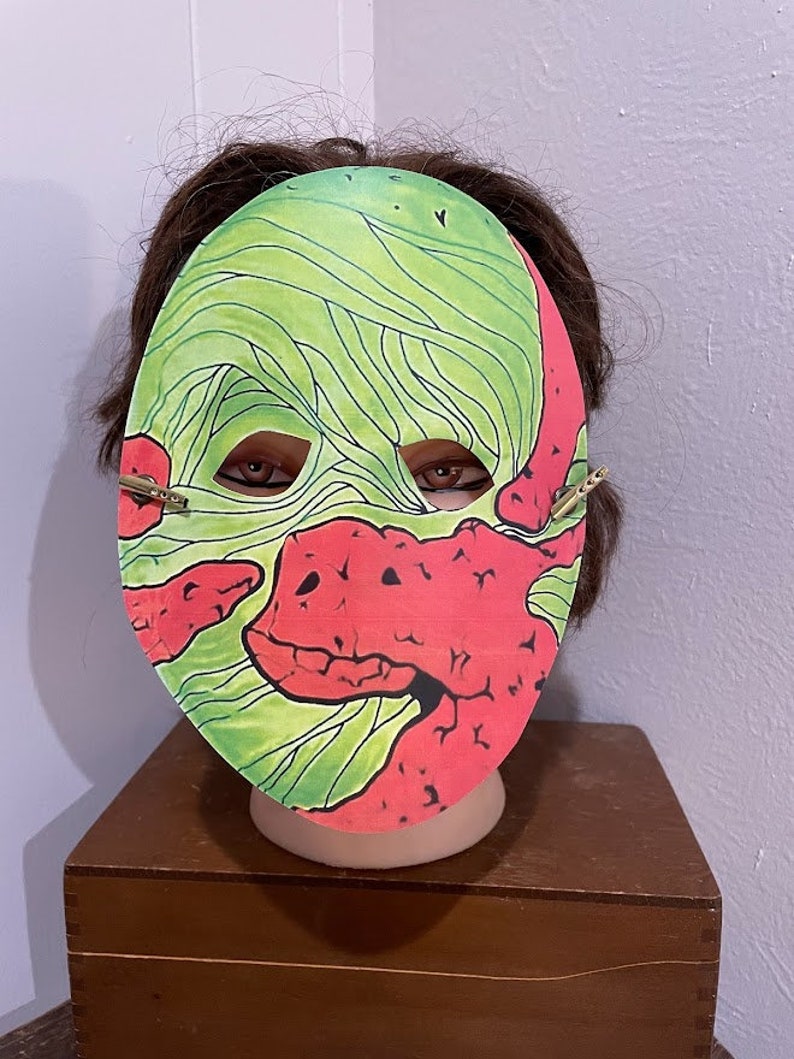 May include: A green and red paper mask with a monster design. The mask has a large mouth and eyes.