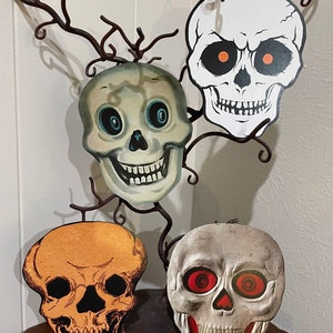 May include: Four Halloween skull decorations made of cardboard. The skulls are white, orange, and gray with black details. The skulls have different facial expressions and eye details.