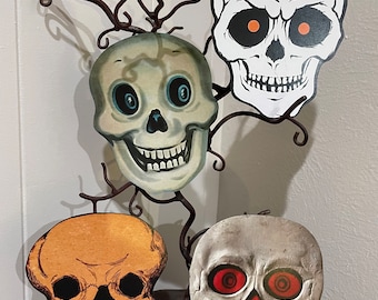 Könnte beinhalten: Vier Halloween-Schädeldekorationen aus Pappe. Die Schädel sind weiß, orange und grau mit schwarzen Details. Die Schädel haben unterschiedliche Gesichtsausdrücke und Augendetails.