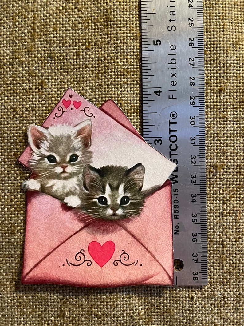 以下が含まれることがあります： 赤いハートと、のぞき込む2匹の子猫がついたピンクの封筒。封筒には、渦巻きとハートの装飾的な縁取りが施されています。子猫は白とグレーで、黒い縞模様をしています。