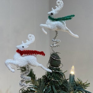 May include: Two white, sparkly reindeer Christmas tree toppers with red and green pipe cleaner scarves. The reindeer are attached to a silver spring.