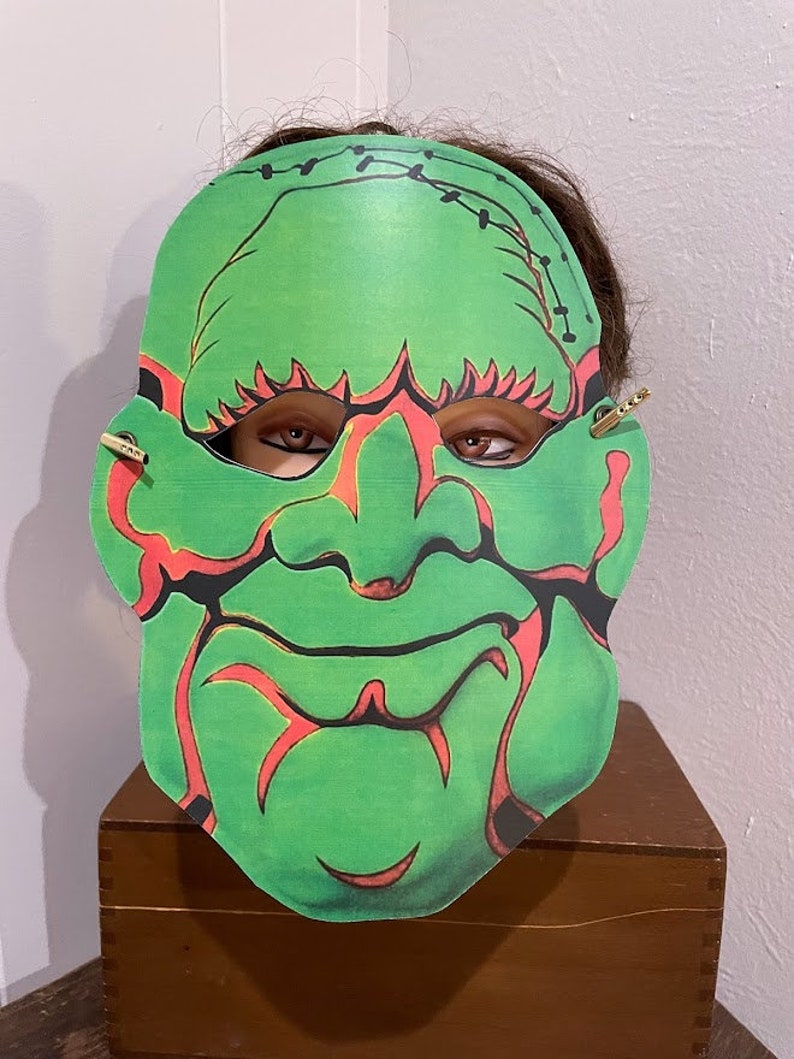 May include: Green monster mask with red stitching and eyes. The mask is made of paper and has a stitched-up look.