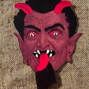 May include: A red devil mask with black hair, red horns, and a red tongue sticking out. The devil's eyes are white with black pupils.