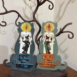 May include: Two decorative candle holders shaped like cats with a witch and a broom inside the candle. The candle holders are made of paper and have a blue and orange color scheme.