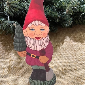 May include: A red and white gnome figurine holding a small green tree and a shovel. The gnome is wearing a red hat, a white beard, and a burgundy shirt with a brown belt.