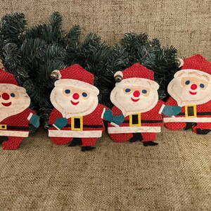 May include: Four vintage Christmas decorations shaped like Father Christmas. The decorations are made of red and white felt with black and yellow details. Each Father Christmas has a white beard and a blue and white hat.