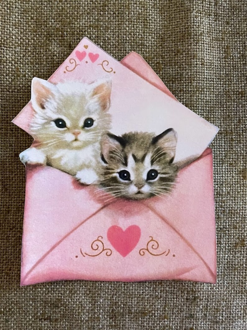 以下が含まれることがあります： ピンクの封筒から顔を出している2匹の子猫。封筒にはピンクのハートと金の渦巻き模様が描かれている。