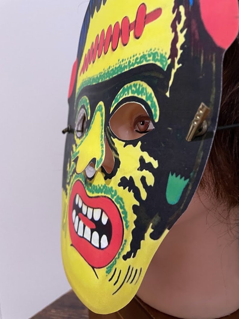 May include: A yellow, green, black and red paper mask of Frankenstein's monster with a stitched forehead and a wide open mouth with teeth. The mask has eye holes and is attached to a string.