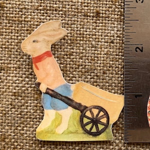 May include: A vintage paper cutout of a white rabbit wearing a red scarf and blue pants pushing a wheelbarrow. The rabbit is on a green background.