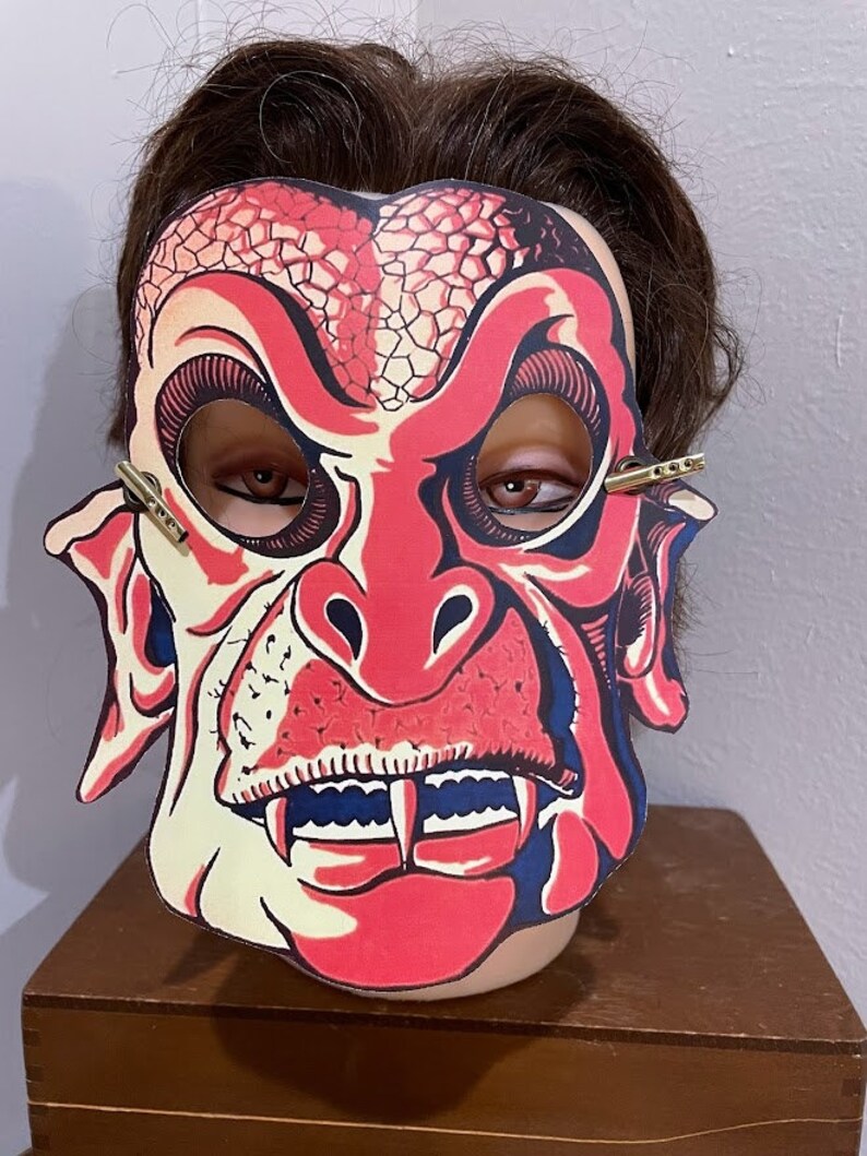 May include: A red and white monster mask with fangs and a textured nose. The mask has eye holes and is attached to a headband.