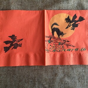 May include: Orange paper napkins with a Halloween theme. The napkins feature a black cat, two witches flying on broomsticks, and two jack-o'-lanterns. The background is a bright orange with a yellow moon.
