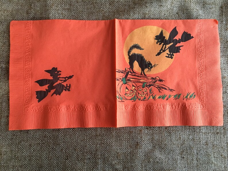 May include: Orange paper napkins with a Halloween theme. The napkins feature a black cat, two witches flying on broomsticks, and two jack-o'-lanterns. The background is a bright orange with a yellow moon.
