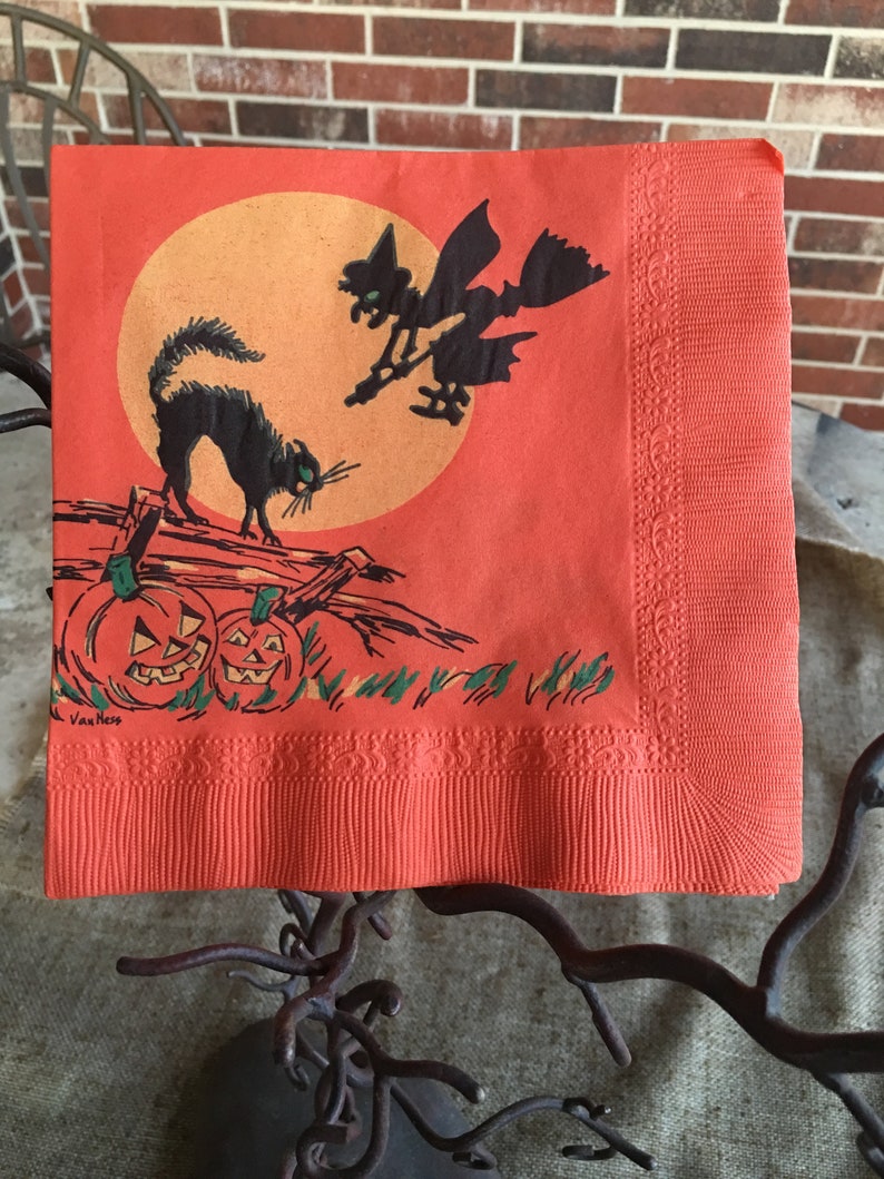 May include: Orange paper napkin with a black cat, a witch flying on a broomstick, and two jack-o'-lanterns. The background is a large orange circle with a yellow center. The text "Van Ness" is printed in the bottom left corner.