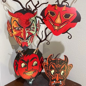 Puede incluir: Cuatro máscaras de diablo vintage de Halloween, cada una con una expresión diferente, se muestran en una rama de metal negro. Las máscaras están hechas de papel y tienen colores rojo, naranja y negro.