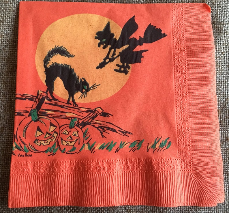 May include: Orange paper napkin with a Halloween theme. The napkin features a black cat, a witch flying on a broomstick, and two jack-o'-lanterns. The background is a large orange moon with a yellow center. The napkin is decorated with a scalloped border.