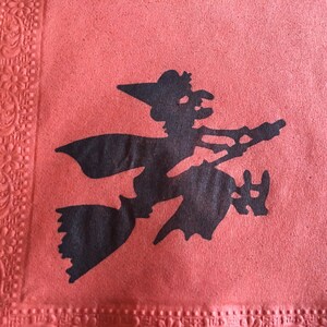 May include: A black silhouette of a witch flying on a broomstick against a red background. The witch is wearing a pointed hat and has a long flowing cape.
