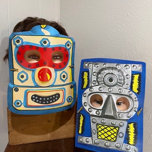 Puede incluir: Dos máscaras de Halloween vintage de robot. La máscara de la izquierda es azul, amarilla y roja con una cara sonriente. La máscara de la derecha es azul, plateada y amarilla con una expresión más seria.