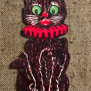 May include: A black cat with green eyes and a red ruffled collar. The cat is sitting with its paws tucked under its body.