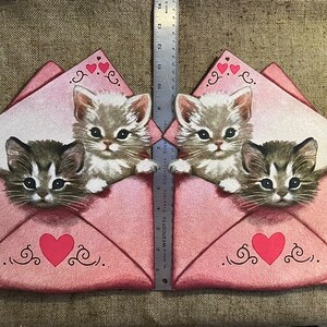 以下が含まれることがあります： ハートと渦巻きが描かれたピンクの封筒が2つ。それぞれの封筒には、上部に顔を出した子猫と、下部に顔を出した大きな子猫がいます。子猫は白地に灰色の模様で、黒い目を持っています。