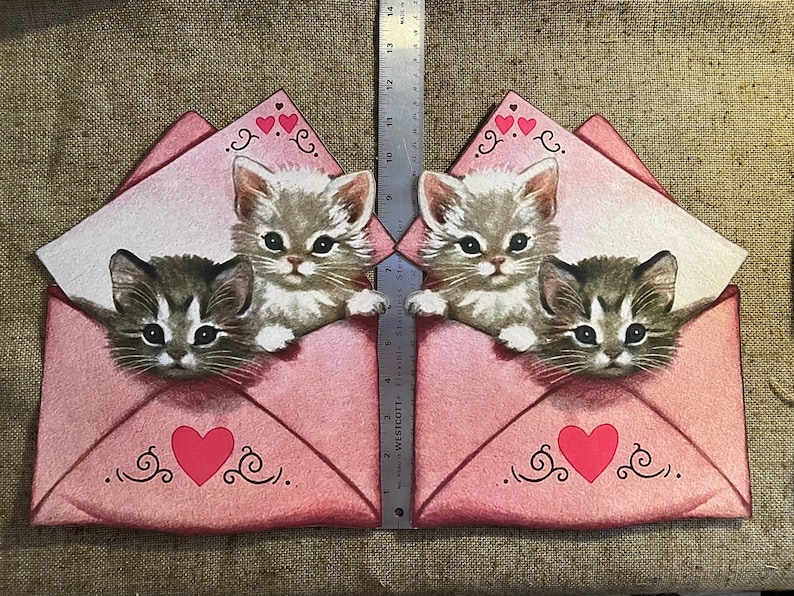 以下が含まれることがあります： ハートと渦巻きが描かれたピンクの封筒が2つ。それぞれの封筒には、上部に顔を出した子猫と、下部に顔を出した大きな子猫がいます。子猫は白地に灰色の模様で、黒い目を持っています。