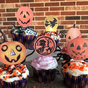 May include: Six Halloween-themed cupcakes decorated with orange and black sprinkles. Each cupcake has a different pumpkin-shaped paper topper with a jack-o'-lantern face. One topper says "Treat".