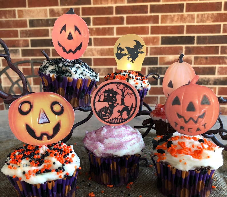 May include: Six Halloween-themed cupcakes decorated with orange and black sprinkles. Each cupcake has a different pumpkin-shaped paper topper with a jack-o'-lantern face. One topper says "Treat".
