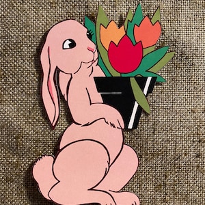 May include: A pink bunny rabbit holding a bouquet of three tulips in a black pot. The tulips are red, orange, and yellow.