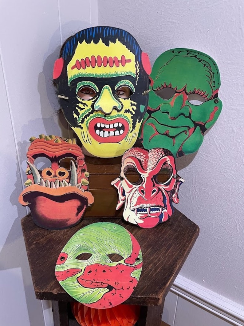 May include: Five vintage Halloween masks featuring monster faces. The masks are made of cardboard and have a colourful design. The masks are green, red, yellow, and black.