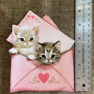 以下が含まれることがあります： ハートが描かれたピンクの封筒。白い子猫と虎柄の子猫が封筒から顔を出しています。