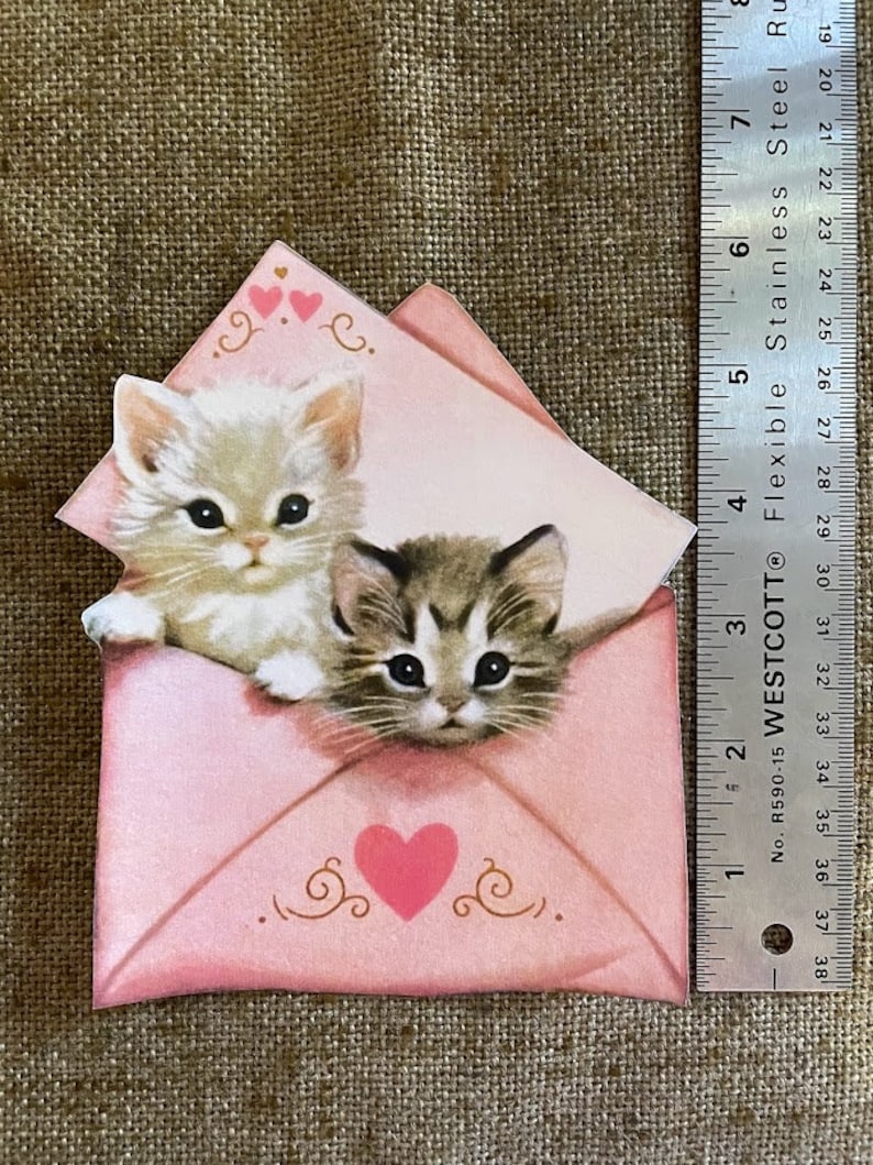 以下が含まれることがあります： ハートが描かれたピンクの封筒。白い子猫と虎柄の子猫が封筒から顔を出しています。