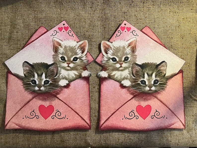 以下が含まれることがあります： 白い紙がのぞいているピンクの封筒が2つあり、それぞれに子猫が2匹ずつ入っています。1匹の子猫は白地に灰色の模様、もう1匹は灰色地に白い模様をしています。両方の封筒には赤いハートと装飾的な渦巻き模様があります。